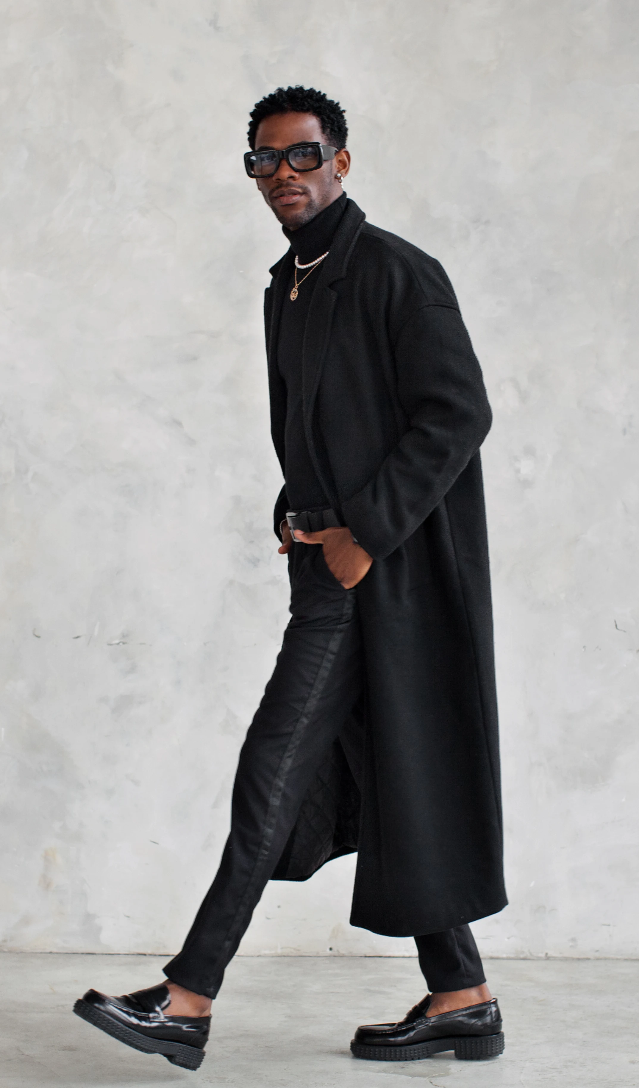 Person wearing a long black coat and black pants against a gray background.