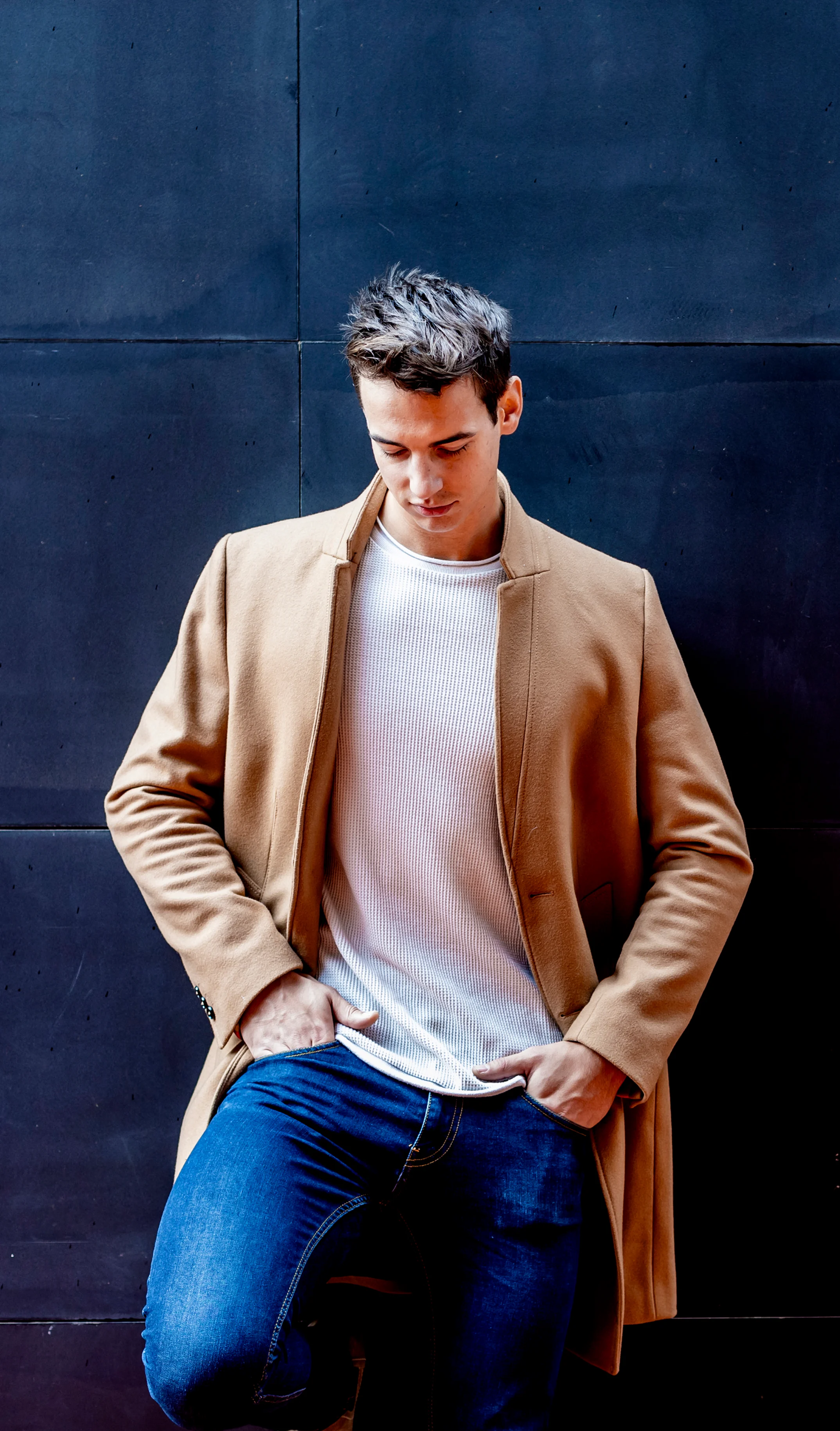 A man in a beige coat and white shirt poses confidently against a dark background.