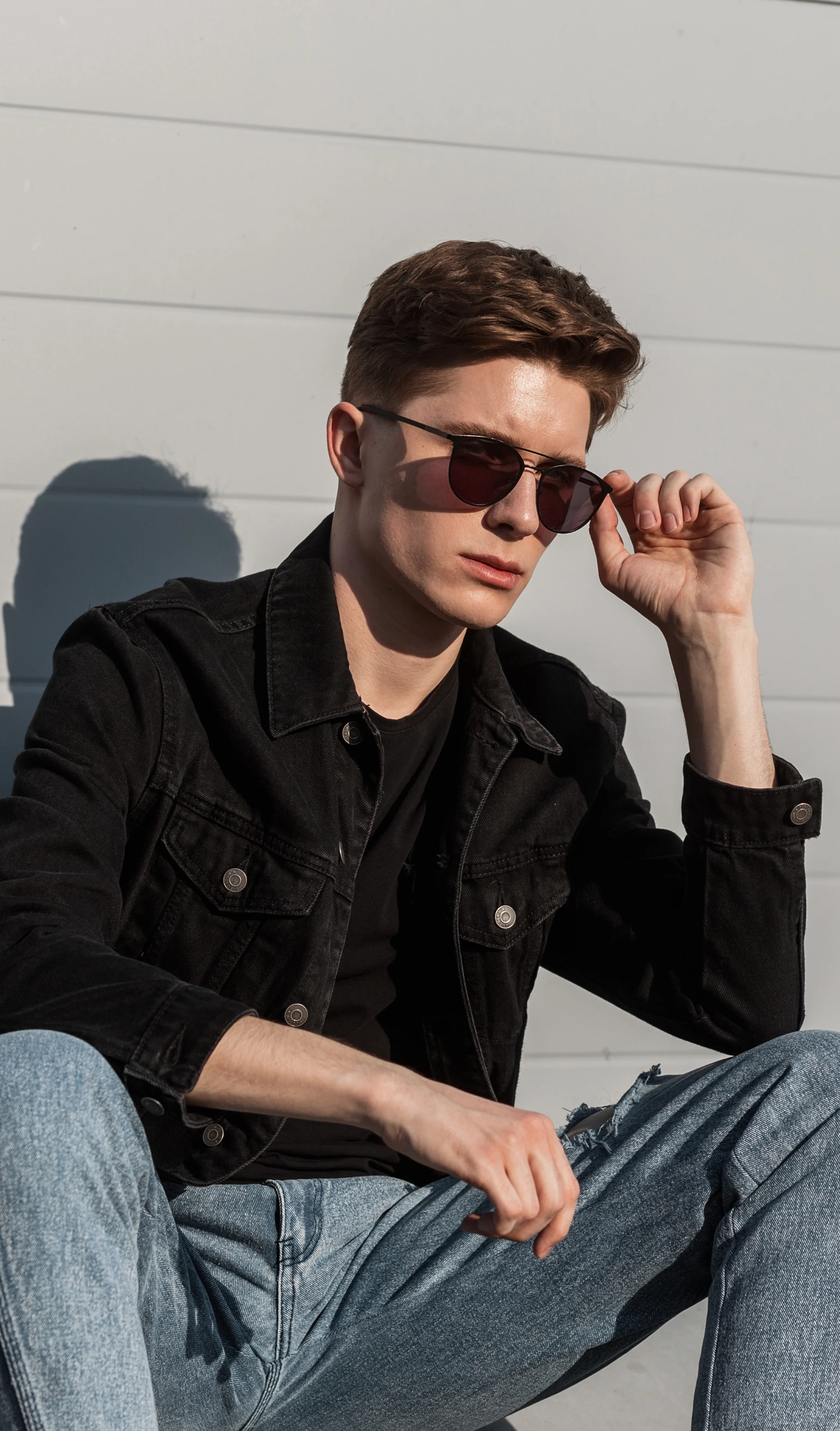 Young man in sunglasses adjusting them while sitting outdoors.