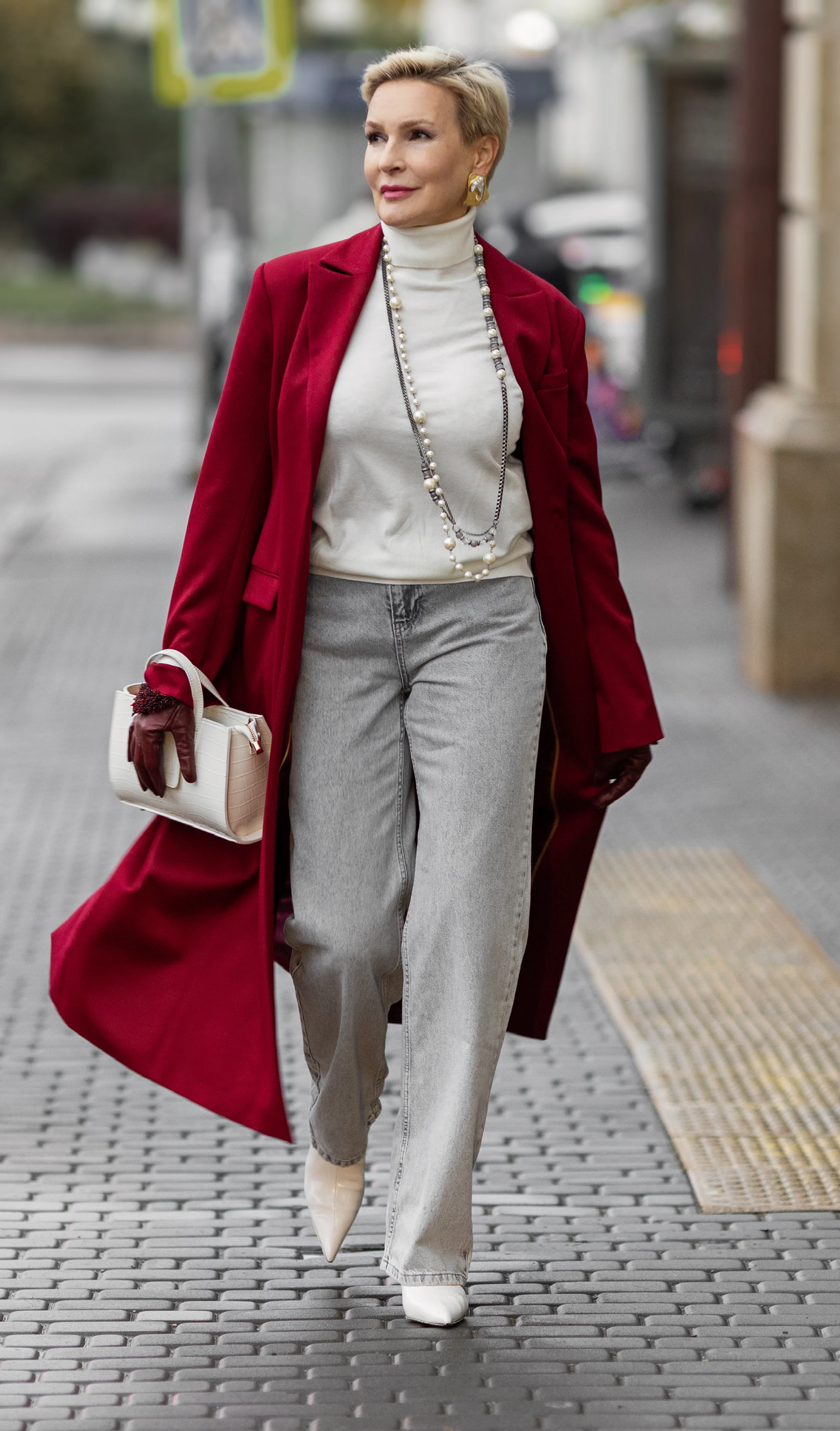 Woman walking in stylish red coat and light pants, holding a small white handbag.