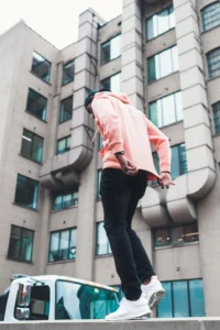 Person in pink hoodie jumping near a building with large pipes.