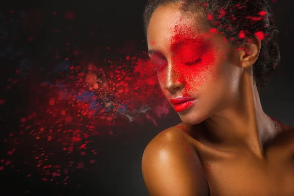 Woman with eyes closed, vibrant red and orange powder on face and shoulder.
