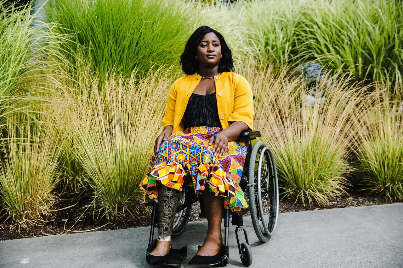 Woman in a wheelchair wearing a colorful skirt and yellow jacket outdoors.