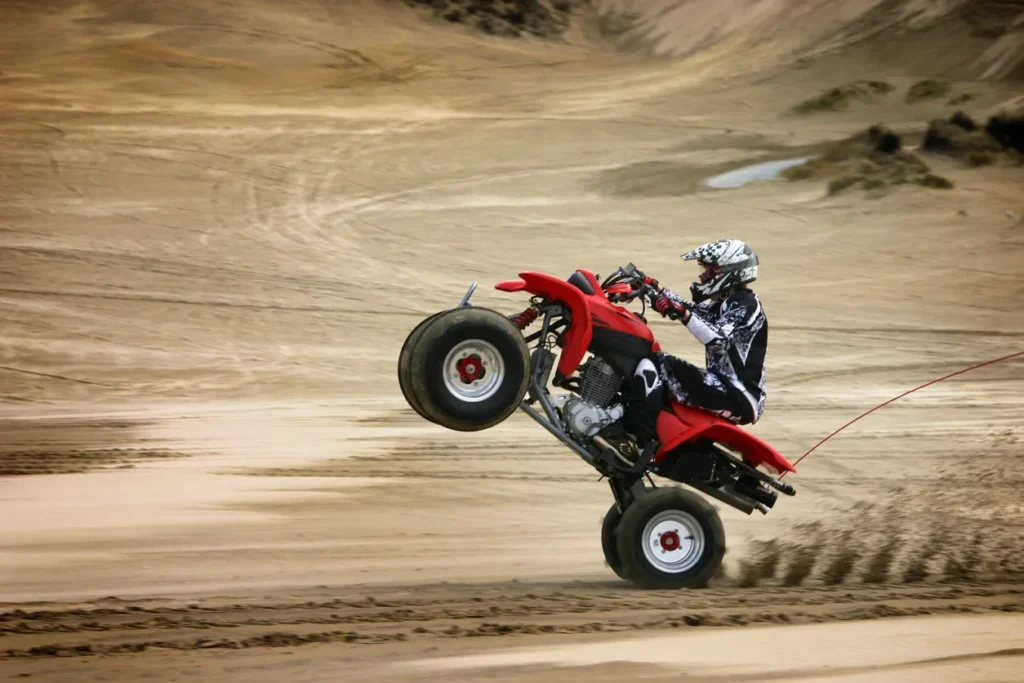 Person performing a wheelie on a red ATV in a sandy area.