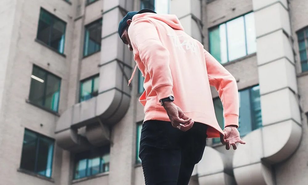 Person in a pink hoodie stretching arms outdoors in urban setting.
