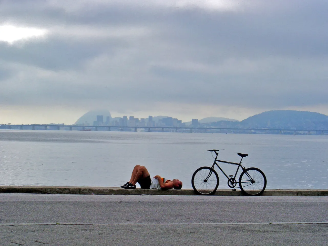 Person lying on the ground next to a bicycle by the waterfront under a cloudy sky.