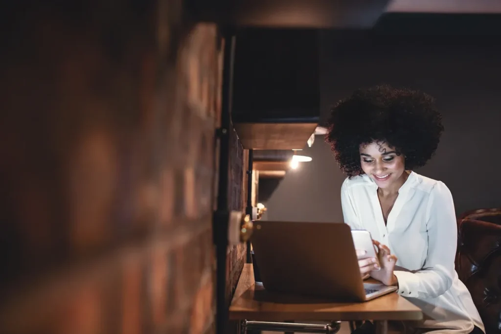 Woman smiling and working on a laptop in a cozy dimly lit space.