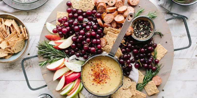 Cheese fondue with apples, grapes, bread, and seasoning on a wooden board.